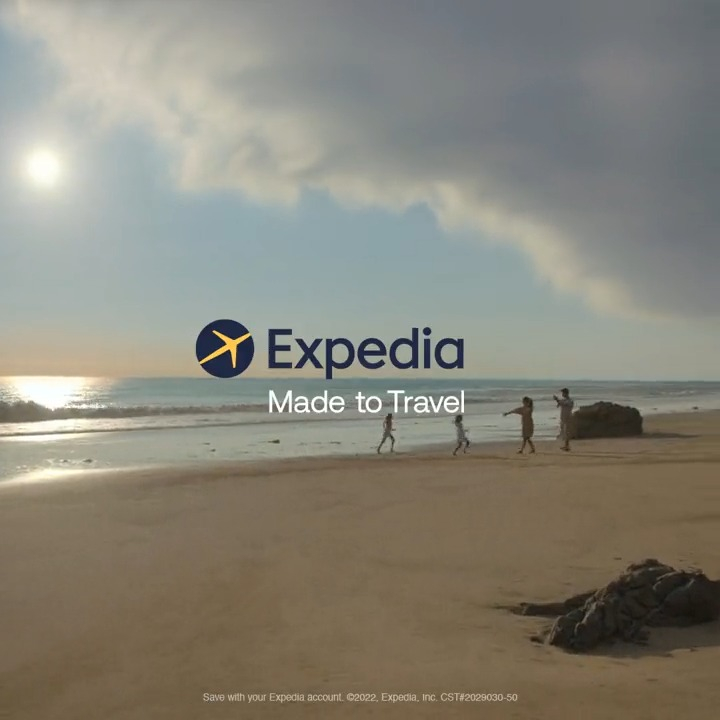 A scenic beach view featuring a group of people walking along the shore with a bright sun shining and soft clouds in the sky. The Expedia logo and the tagline 'Made to Travel' are prominently displayed.